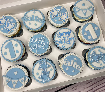 Box of cupcakes with blue and white decorations, including stars and the number 1