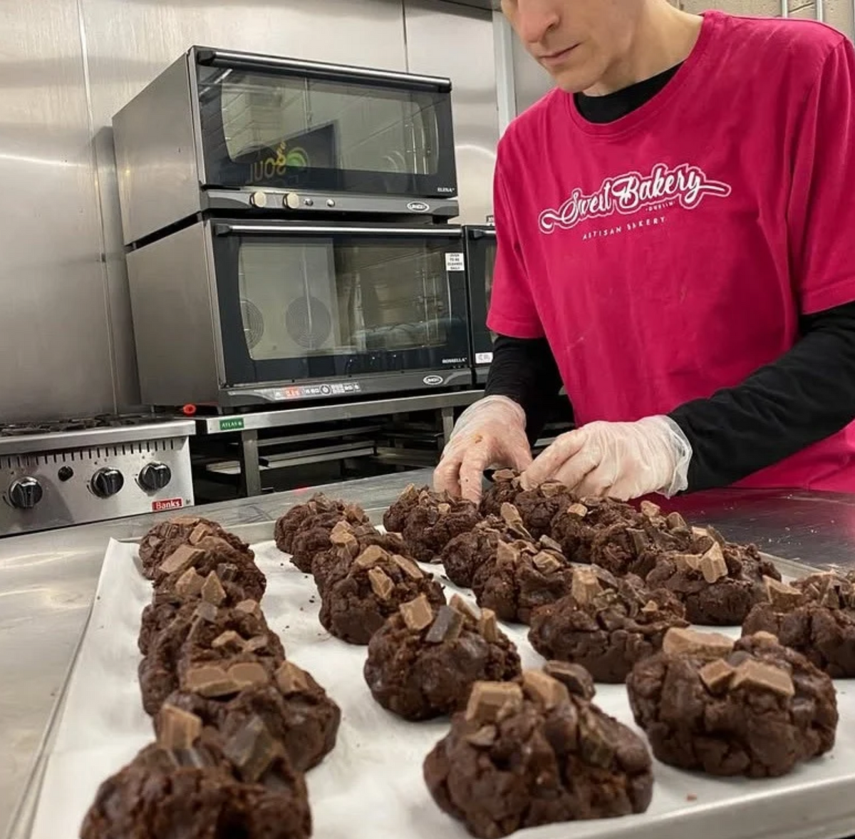 A person in a bright pink Bakery t-shirt and white gloves arranges chocolate chip cookies on a baking sheet in a commercial kitchen.